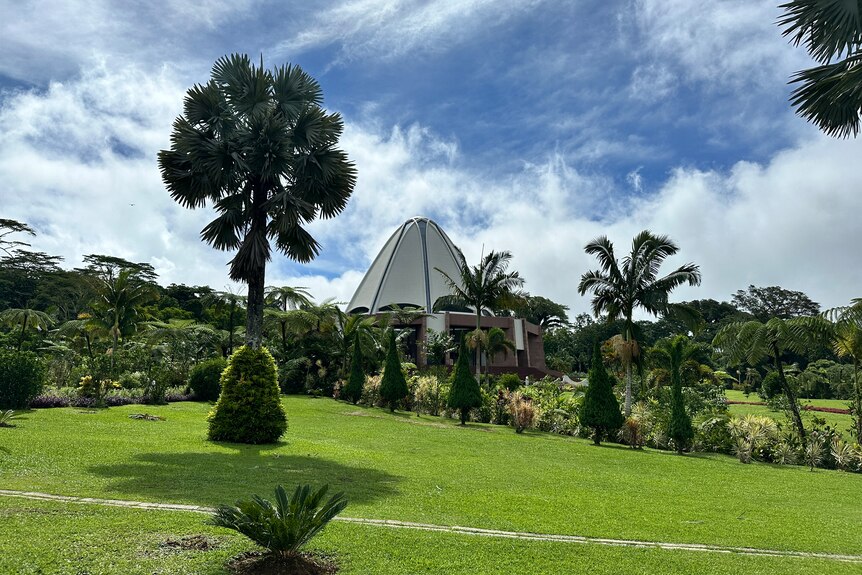 Samoa's Baha'i Temple, a white dome shaped building, under blue skies with some clouds, and green grass and palm trees in front.