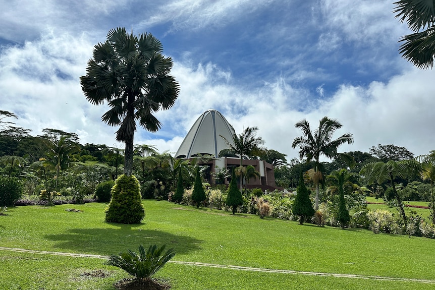 Samoa's Baha'i Temple, a white dome shaped building, under blue skies with some clouds, and green grass and palm trees in front.
