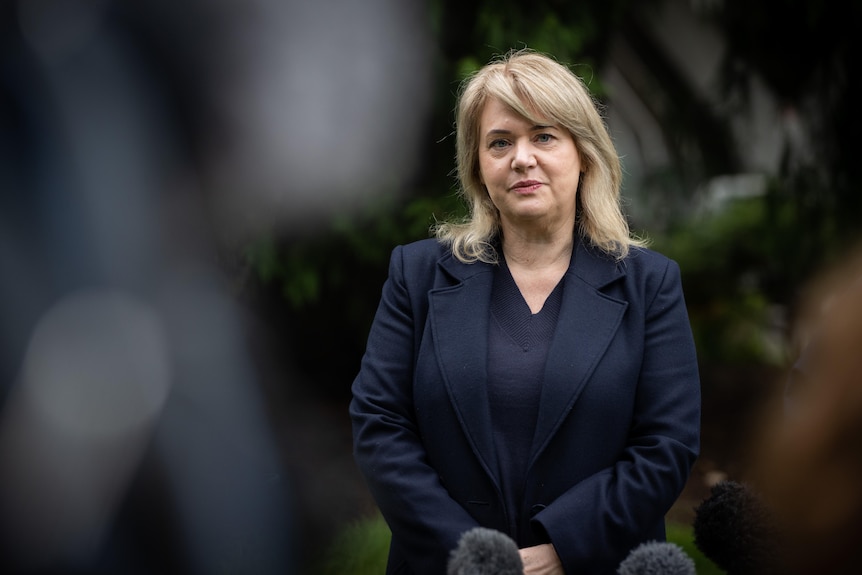 A woman with blonde hair wearing a dark suits looks at the camera