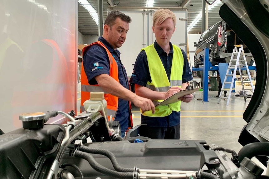 Two men wearing high-vis vests inspect the engine or a truck.