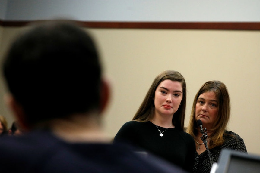 A tearful teen speaks at a microphone as she stares at a man, seen in the foreground of the image.