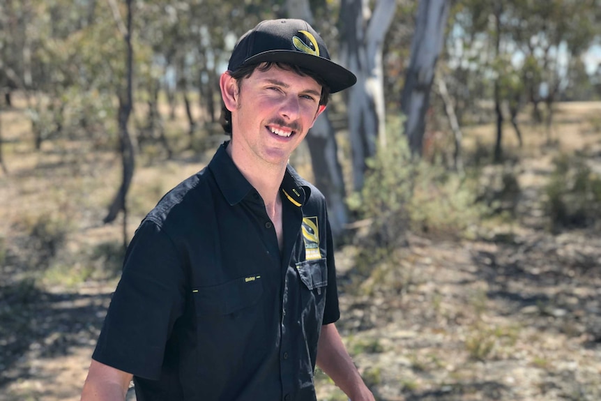 Canberra snake catcher Luke Dunn stands in the bush, smiling and wearing and hat.