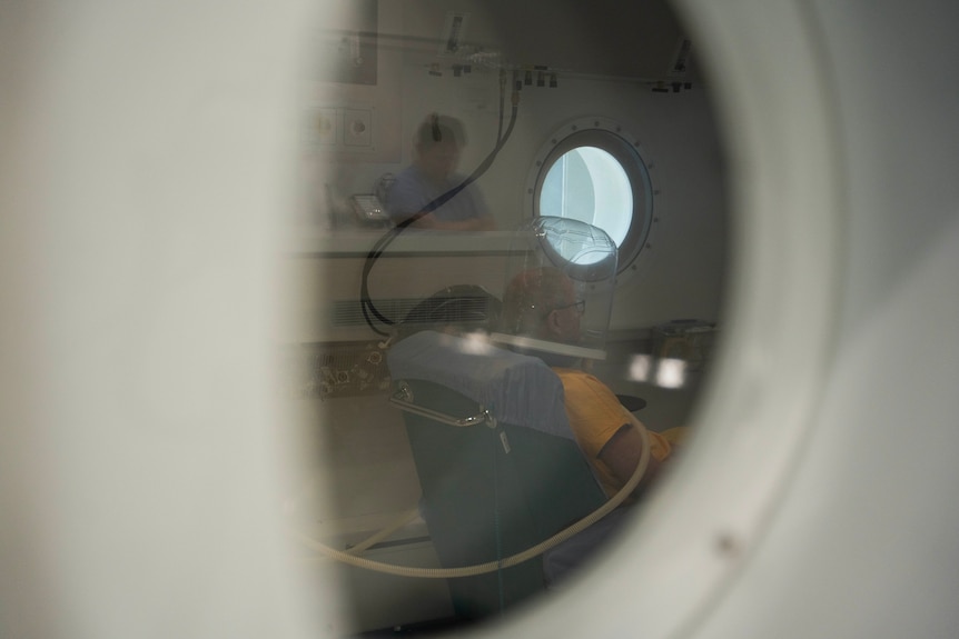 Looking through a round window in a hospital ward, a person sits on a chair with an astronaut-like clear head piece on.