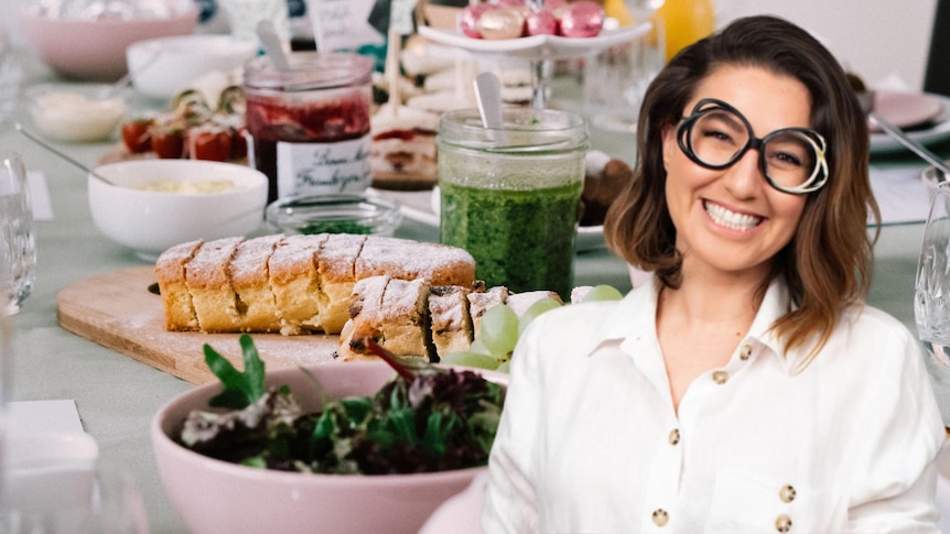 A woman with glasses is smiling, in front of another photo of a table filled with food and drinks. 