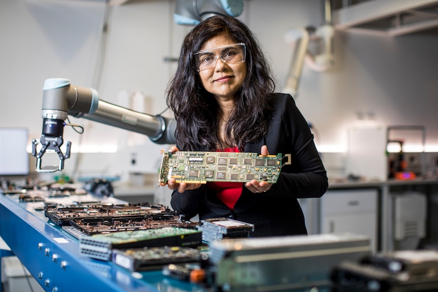 a woman in goggles in a lab holding up a circuit board