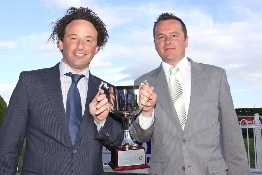 Two men in suits hold up a racing trophy together.