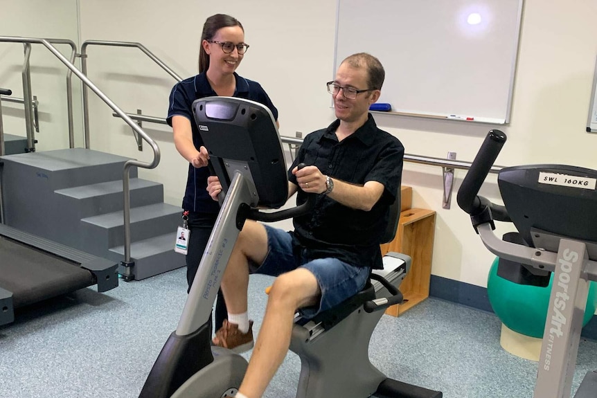 Joshua Leveridge rides a stationary exercise bike in hospital with a therapist.
