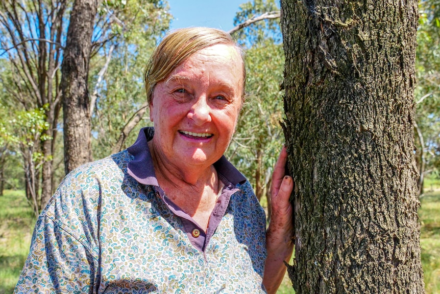 An older woman stands beside a tree in a paddock