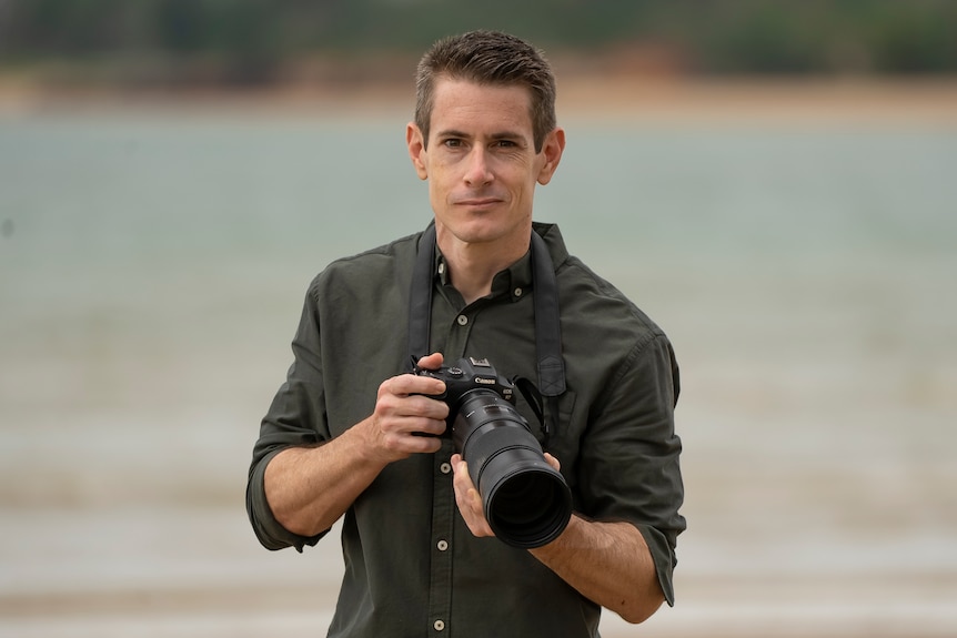 Brandon Sideleau stands in front of a body of water holding a camera.