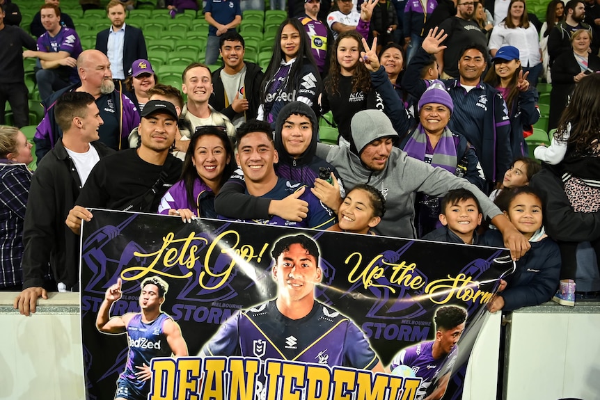 Dean Ieremia celebrates with a Melbourne Storm banner, surrounded by hugging family and friends.