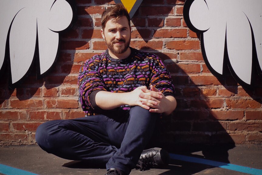 A man wearing bright spotted jumper and jeans sits leaning against a brick wall under artwork