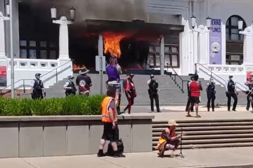 Police stand outside a building as its door burns.