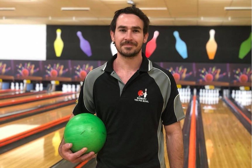 A man holding a ten pin bowling ball with a bowling alley behind him.