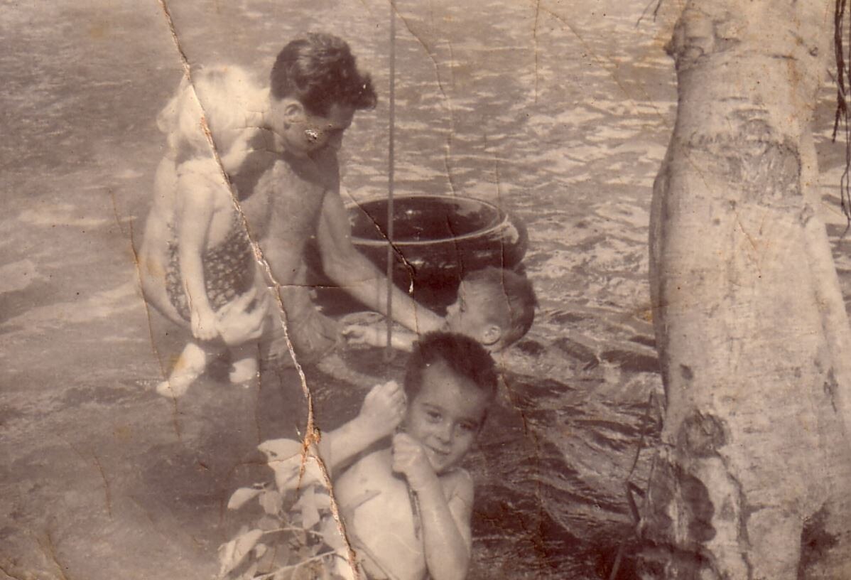 A father swims in a waterhole with his three young children. He is holding his daughter on his hip.