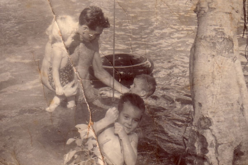 A father swims in a waterhole with his three young children. He is holding his daughter on his hip.