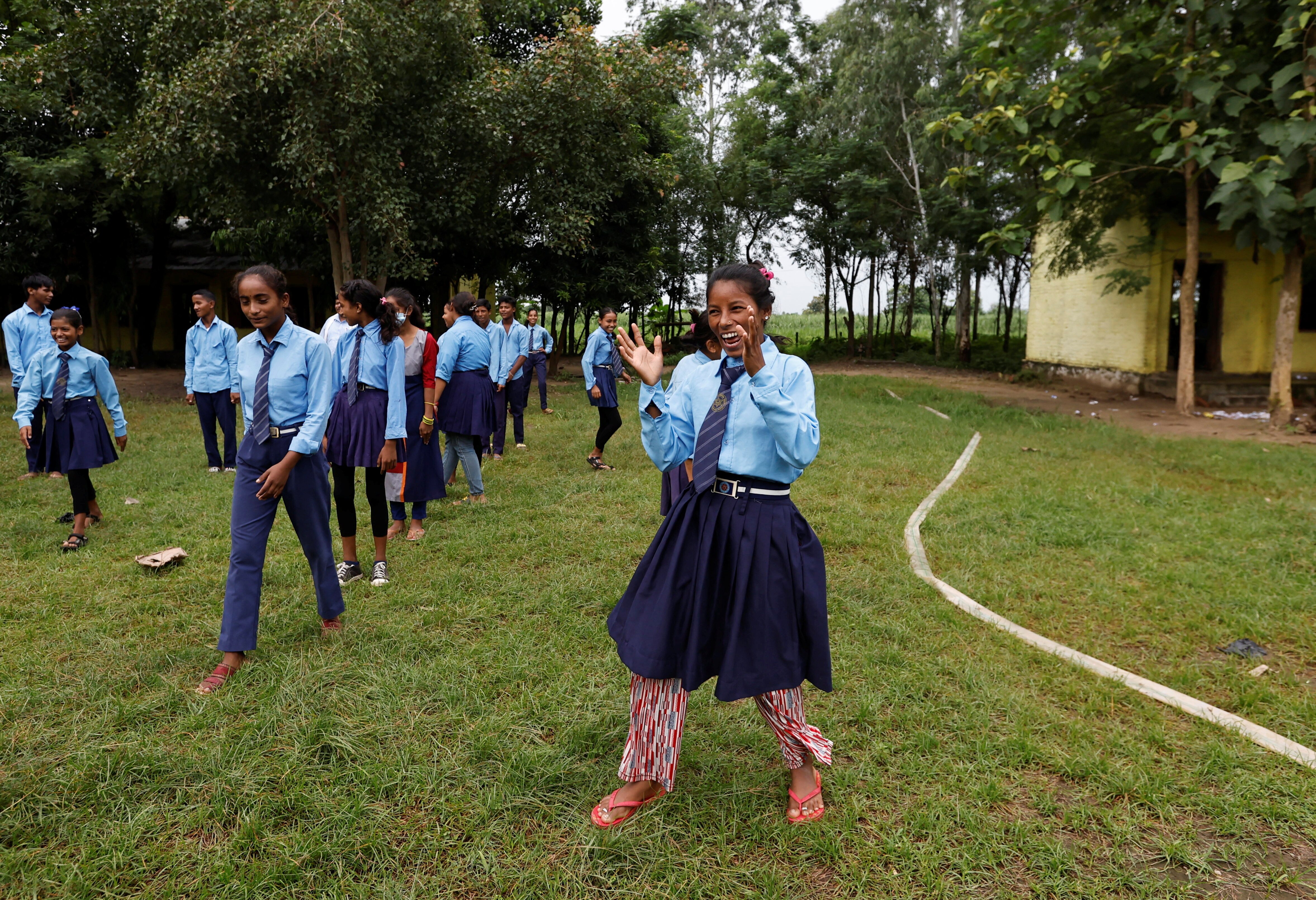 A woman in school uniform stands in a grass yard laughing and clapping her hands. Students walk in a line beside her 