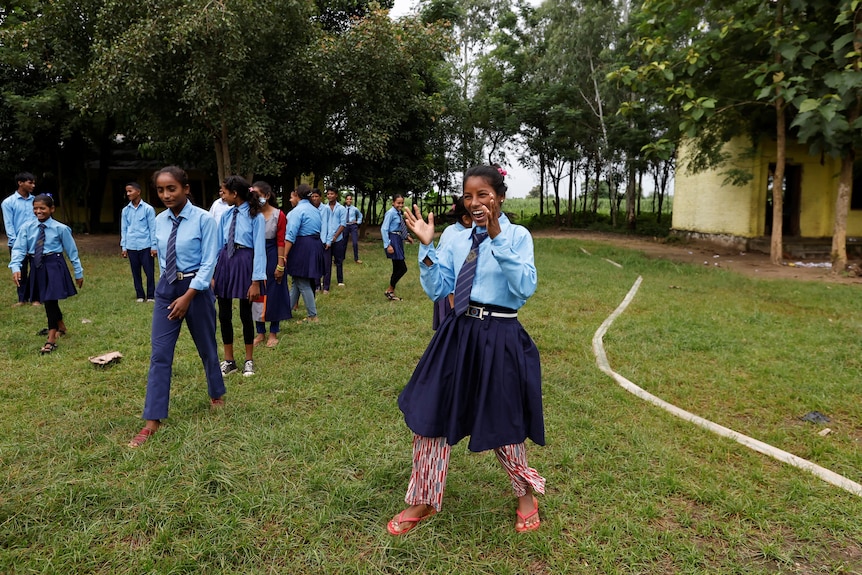 A woman in school uniform stands in a grass yard laughing and clapping her hands. Students walk in a line beside her 