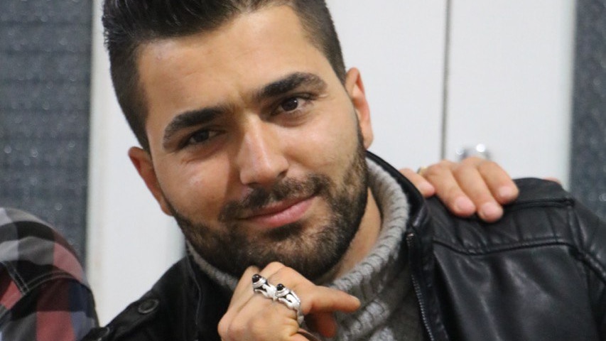 A man with a trimmed beard wearing lots of rings and a leather jacket smiles at camera