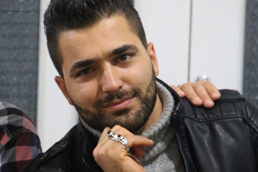 A man with a trimmed beard wearing lots of rings and a leather jacket smiles at camera
