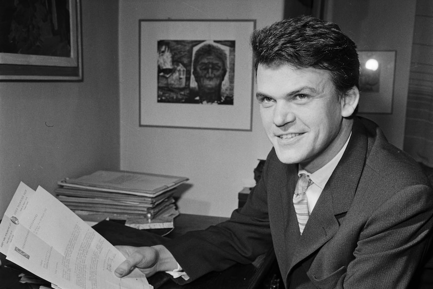 A black and white photo of a young man in a suit holding papers 