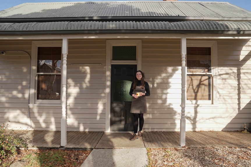 A young woman wearing grey dress over black tights and skivvy stands in front of a classic Australian home