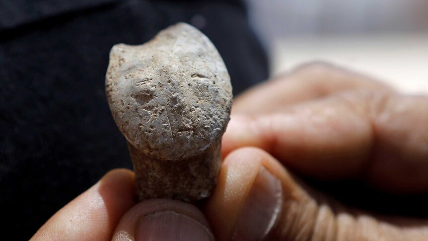 A stone figurine the size of a thumb depicts a human face, which is held close to the camera and held between a person's hands.