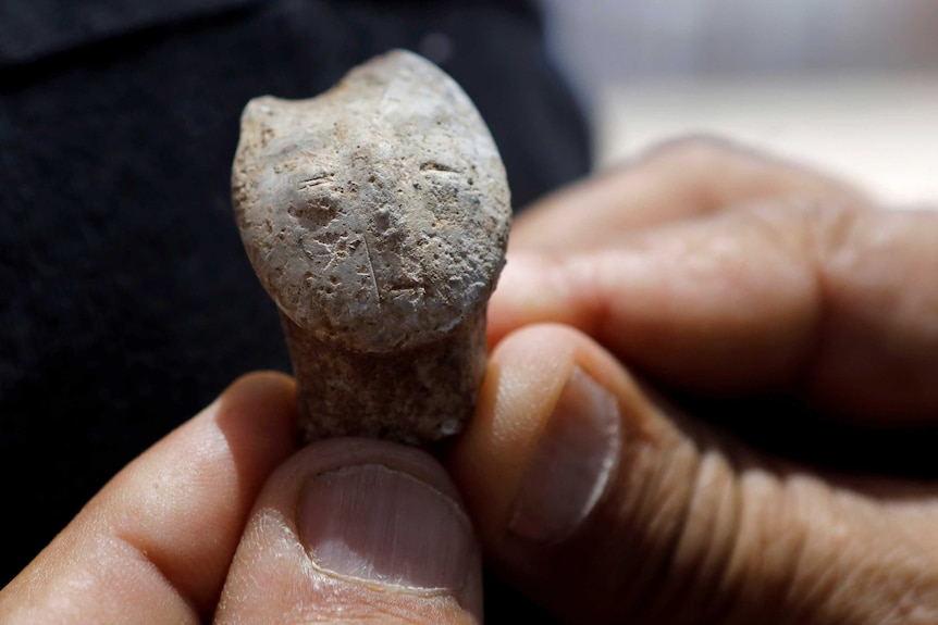 A stone figurine the size of a thumb depicts a human face, which is held close to the camera and held between a person's hands.