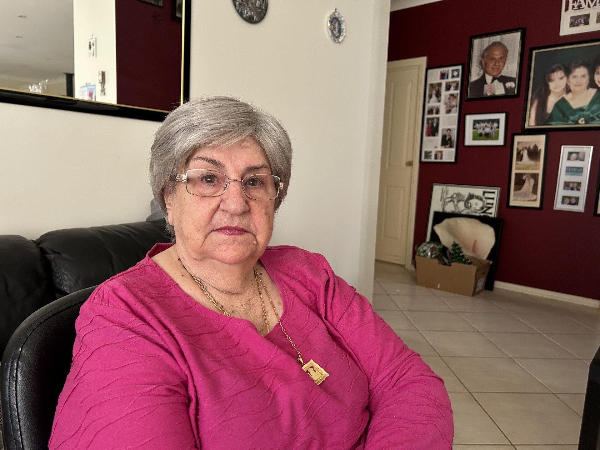 Teresa de cabelos grisalhos e suéter rosa sentada, séria, sala com fotos na parede, uma caixa embaixo.