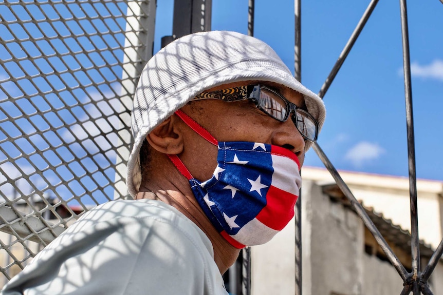 Man in white hat and face mask with American flag leaning on wire fence