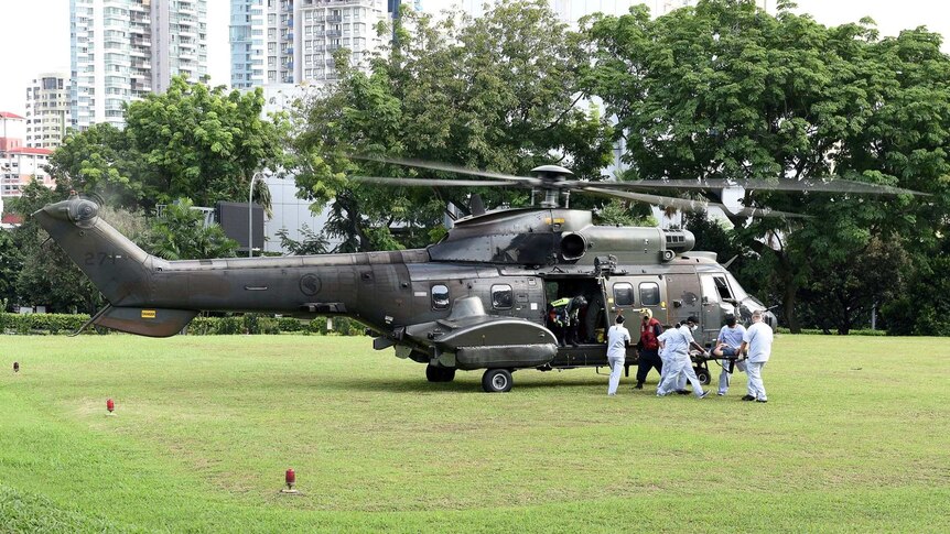 A large military helicopter is parked on a grassy field with people dressed in white carrying somebody on a stretcher.