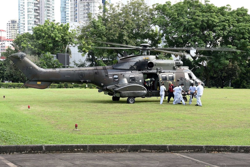 A large military helicopter is parked on a grassy field with people dressed in white carrying somebody on a stretcher.