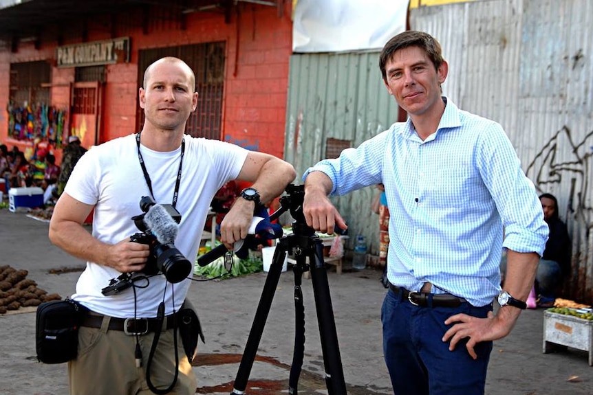 Cochrane holding camera and Tlozek leaning on tripod with corrugated fence in background.