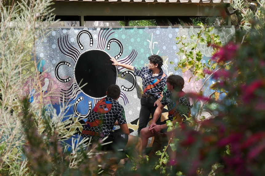 A vignette of out of focus plants with a wide shot of three students sitting in front of an Aboriginal mural.