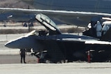 A blurry photograph of a het on a runway. Several people wearing protective suits are standing on and around the aircraft.