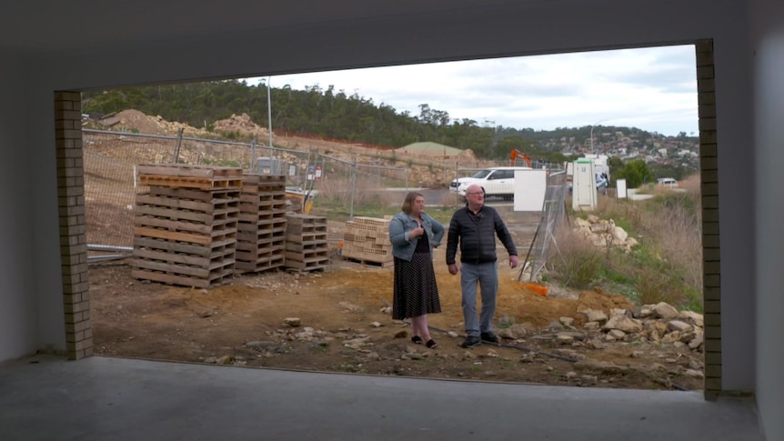 A man and a woman stand outside an open garage door, with construction materials behind them.