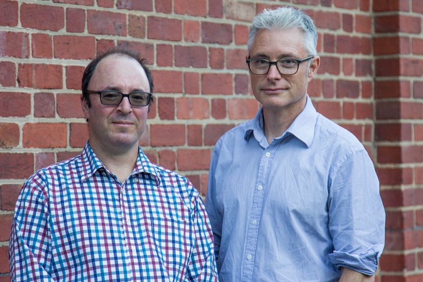 Two bespectacled men stand in front of a brick wall, looking at the camera.