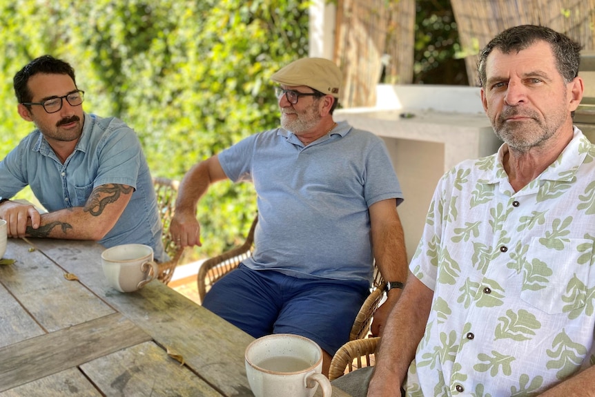 Three men sitting outside at a home bbq area, two are talking in the background and one closest to camera is not smiling