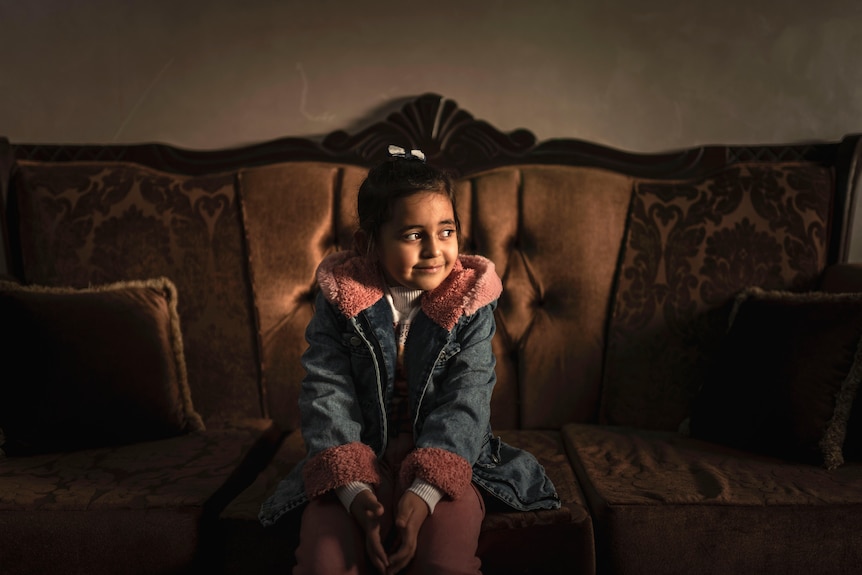 A girl wearing a denim jacket looks away as she sits on a couch.