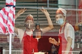 Workers in cafe uniforms and masks are in a food venue. One makes peace signs.