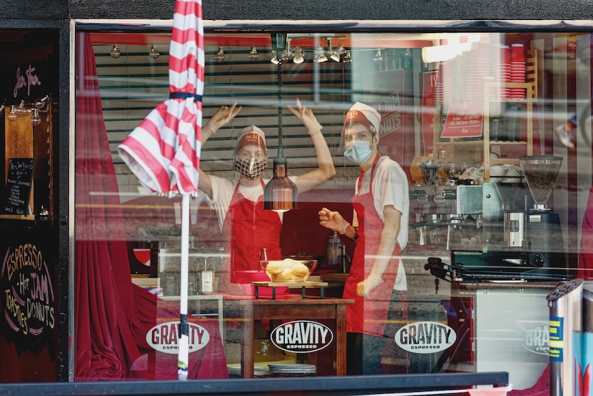 Workers in cafe uniforms and masks are in a food venue. One makes peace signs.