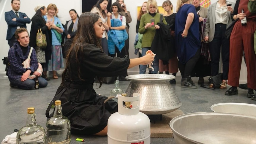 The artist putting rose petals in a pot surrounding by distilling equipment, a crowd watching on.