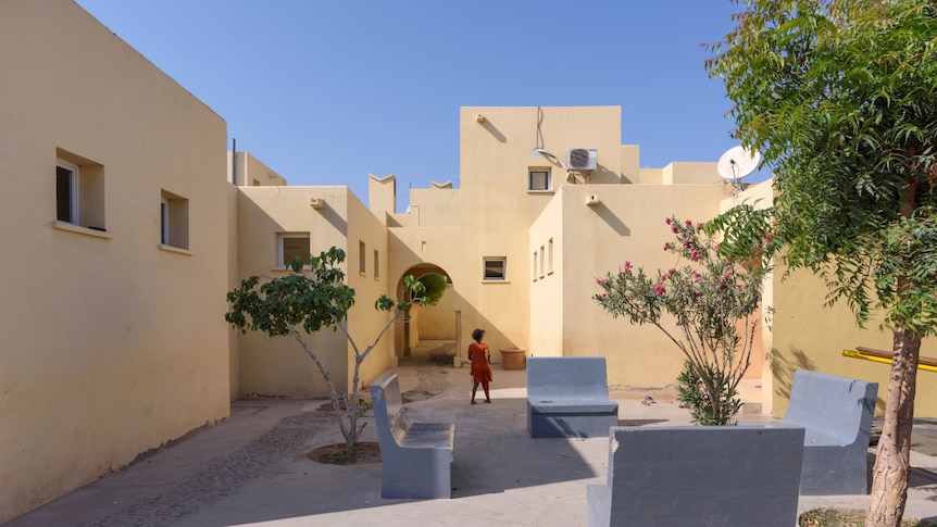 On a clear day, you view a square in a bright yellow medina-style complex with a child facing their back to the camera.