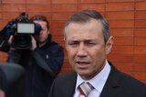 Health Minister Roger Cook standing in front of a brick wall, with a cameraman in the background