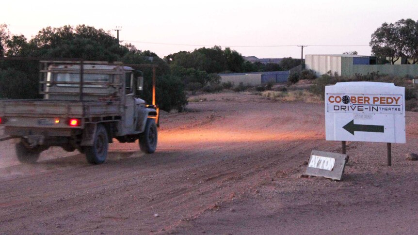 An old ute drives along a sweeping red dirt road past a sign for the Coober Pedy Drive-In at sunset