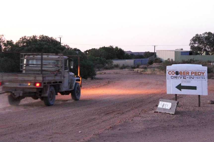 An old ute drives along a sweeping red dirt road past a sign for the Coober Pedy Drive-In at sunset