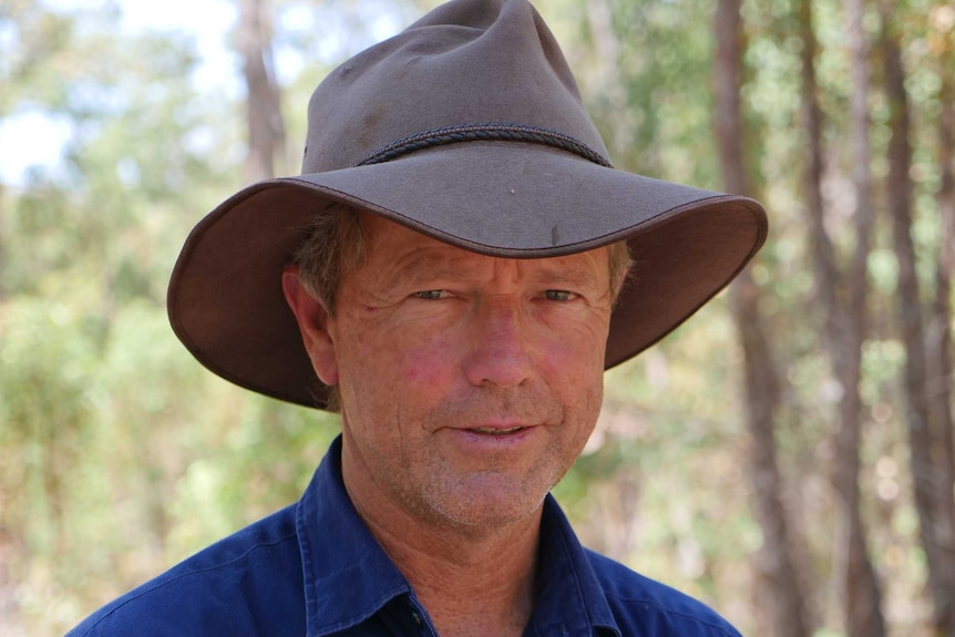 A head and shoulders image of a smiling man in a blue shirt and akubra hat in a forest.