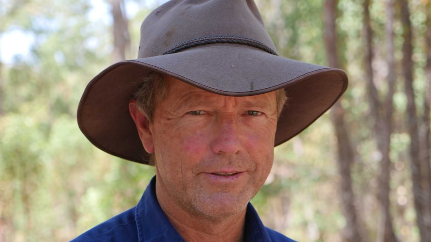 Headshot of a man in a blue shirt and a broad-brimmed hat, standing in a paddock with trees behind him.