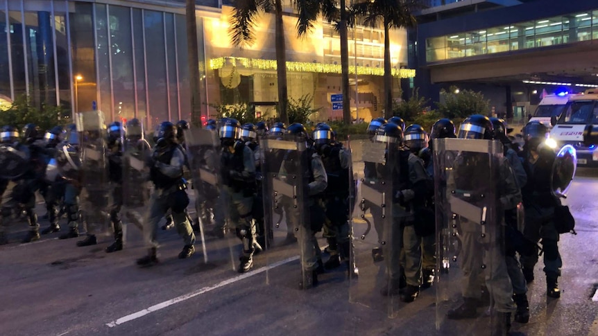 A row of helmeted riot police stand in a street, holding up shields.