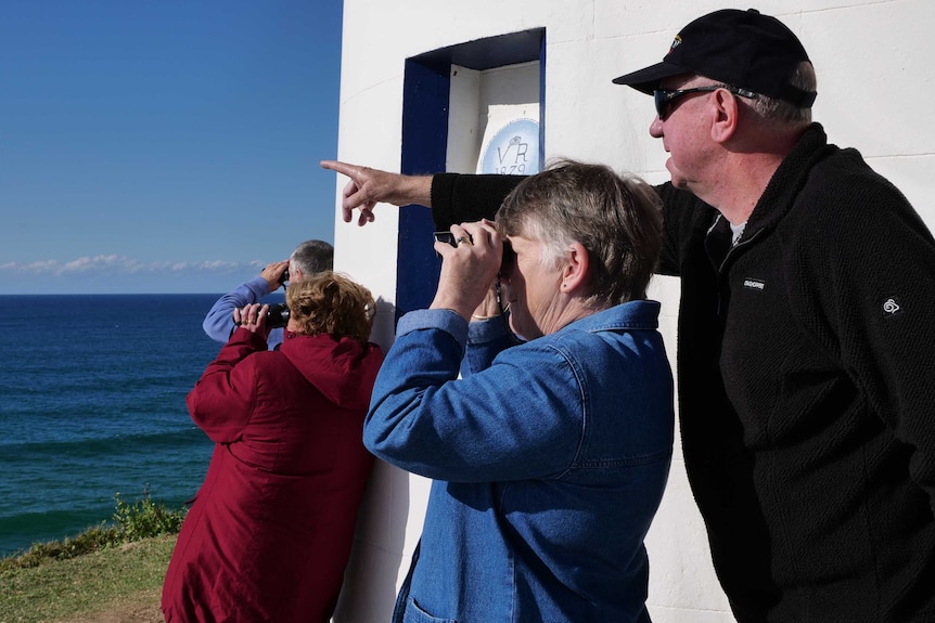 A man and woman stand next to a lighthouse looking and pointing out to the ocean.