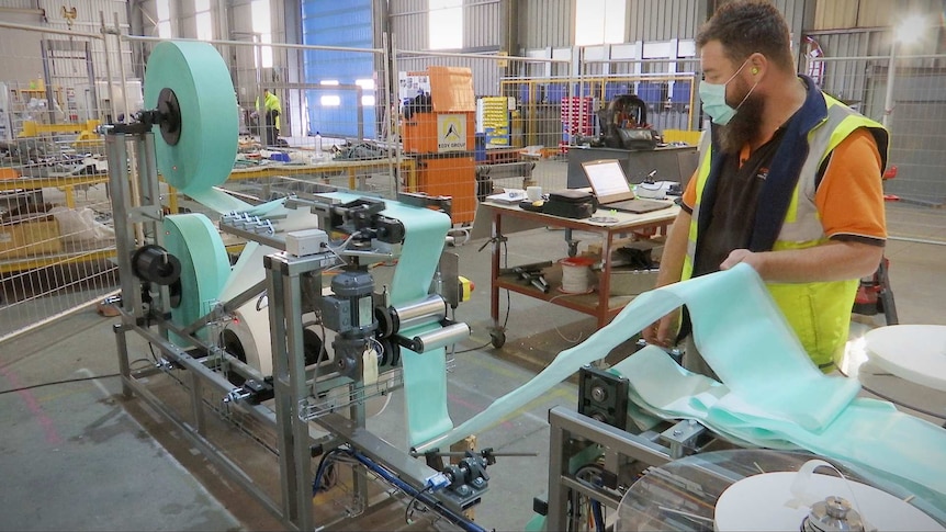 A man in hi-vis work clothing, and surgical mask, holds surgical mask fabric as he watches it feed through a machine.